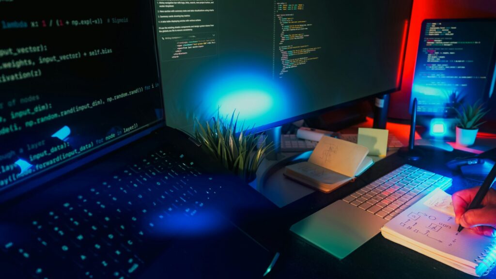 Laptop and monitor displaying code in a modern workspace with neon lighting.